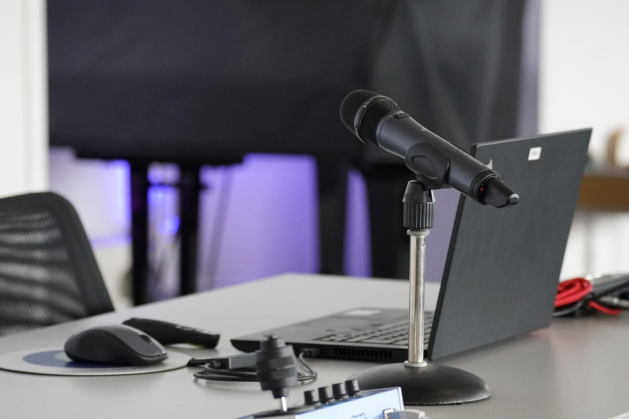 A black microphone on a stand is positioned in front of an open laptop on a desk. Other items, including a computer mouse and audio equipment, are visible on the desk. The background is a blurred room with purple lighting.