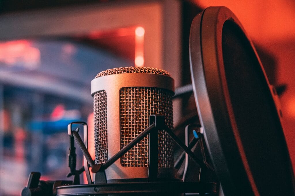 A close-up, angled shot of a gold and black condenser microphone with a pop filter. The background is a warm, out-of-focus scene with red and orange lighting.